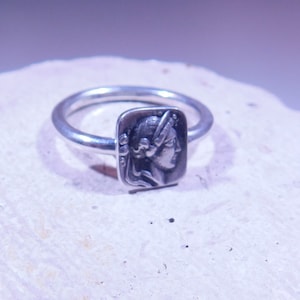 Può includere: Un anello d'argento con un designo quadrato che presenta un'immagine in rilievo del viso di una donna.