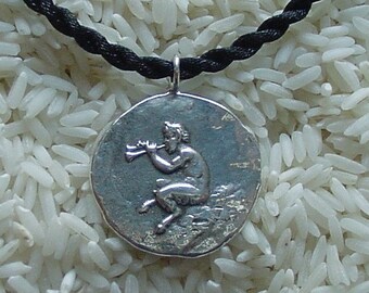 May include: A circular silver-toned pendant featuring a relief of a seated figure playing a horn, suspended from a black braided cord. The pendant has a textured, metallic surface with a raised design of a mythological creature in profile.