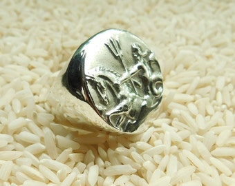 May include: A silver ring with a raised design of a mythical creature holding a trident. The ring is set against a background of white rice.