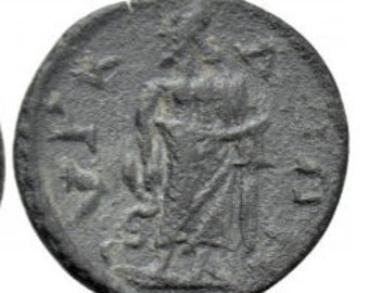 May include: A circular dark grey ancient coin featuring a relief of a standing figure draped in robes. The figure holds a staff-like object in one hand, with faint markings and symbols visible on the textured surface surrounding the central image.