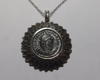 May include: A silver-toned pendant necklace featuring a circular coin with a profile portrait of a figure in relief, surrounded by a decorative border of textured leaves. The pendant hangs from a thin silver-toned chain with a metal bail at the top.