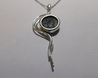 May include: A silver pendant necklace featuring a dark, circular ancient-style coin set within a polished silver frame. The frame extends into a fluid, swirling silver design that tapers downward, suspended from a delicate silver chain.