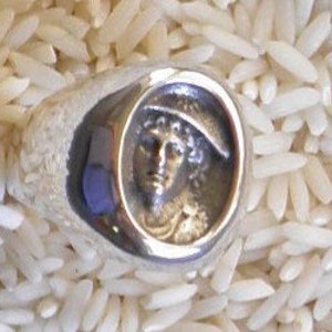 May include: A silver ring with a raised circular design featuring a profile of a person wearing a hat.
