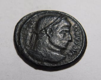 May include: A dark, circular metallic coin featuring a profile portrait of a figure facing right. The surface shows signs of wear and oxidation, with raised relief details depicting facial features and a headband. Faint, weathered lettering is visible.