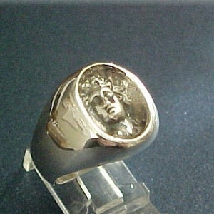 以下が含まれることがあります： 古典的な人物の顔の精巧なレリーフが施されたゴールドカラーのリング。リングは丸みを帯びたシグネットスタイルのデザインです。顔は詳細で質感のあるボーダーで囲まれています。リングは透明なスタンドに展示されています。