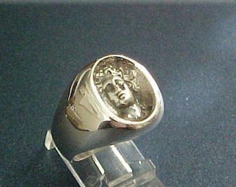 May include: A gold-colored ring with a detailed relief of a classical figure's face. The ring has a rounded, signet-style design. The face is surrounded by a detailed, textured border. The ring is displayed on a clear stand.