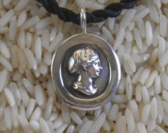 May include: A silver pendant necklace with a round medallion featuring a detailed profile of a woman with a headscarf. The pendant is on a black cord.