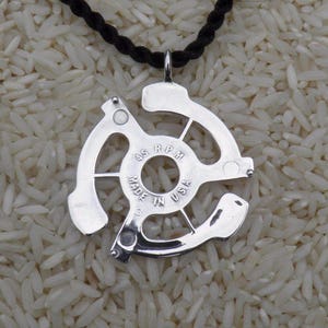 May include: A silver pendant in the shape of a 45 RPM record adapter, suspended from a black cord. The pendant has the words "MADE IN USA" and "45 RPM" inscribed on it. The background is a bed of white rice.
