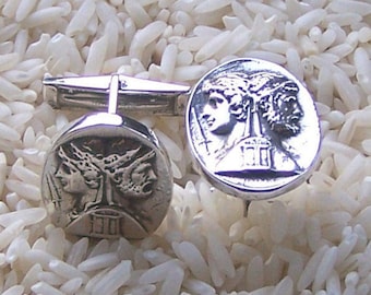 May include: A pair of silver-toned cufflinks featuring a circular face with a relief design of two profile heads facing opposite directions. The design includes a central pillar element and the Roman numeral III at the base of the circular coin-like face.