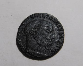 May include: A dark, circular ancient coin featuring a profile portrait of a ruler wearing a laurel wreath. The coin has a textured, weathered surface with raised relief details and Latin text inscribed along the outer edge of the circular border.