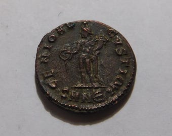 May include: A circular bronze-toned ancient coin featuring a standing figure in the center holding a circular object in one hand and a staff in the other. The coin has raised lettering along the outer edge and a textured, weathered surface.