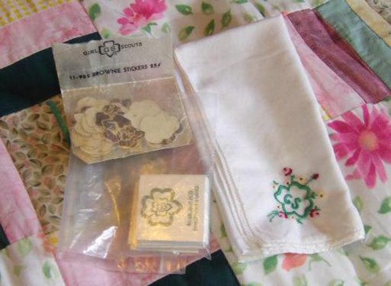 May include: A package of Girl Scout Brownie stickers with a white embroidered handkerchief with the Girl Scout logo and flowers. The package says "Girl Scout Brownie Stickers 35&cent;".