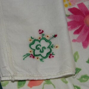 May include: White handkerchief with green embroidered clover and the letters "GS" in the center. The clover is surrounded by small pink and yellow flowers.