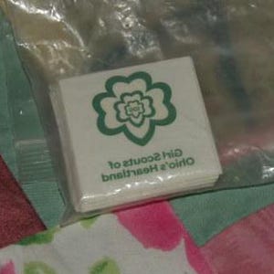 May include: A white package of Girl Scout cookie patches with a green four-leaf clover logo. The text on the package reads "Ohio's Heartland Girl Scouts of".
