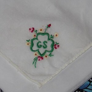May include: White cotton handkerchief with green embroidered four-leaf clover with the letters "GS" in the center. The clover is surrounded by small pink and yellow embroidered flowers.