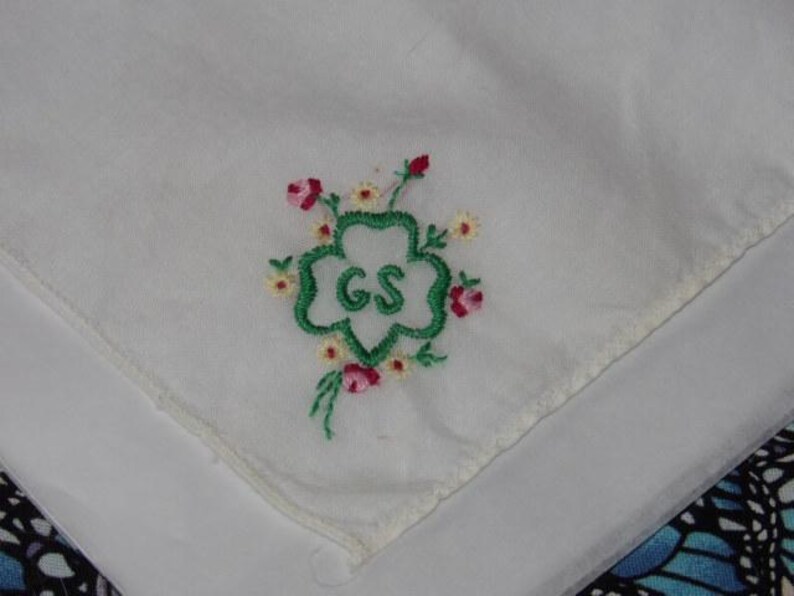 May include: White cotton handkerchief with green embroidered four-leaf clover with the letters "GS" in the center. The clover is surrounded by small pink and yellow embroidered flowers.