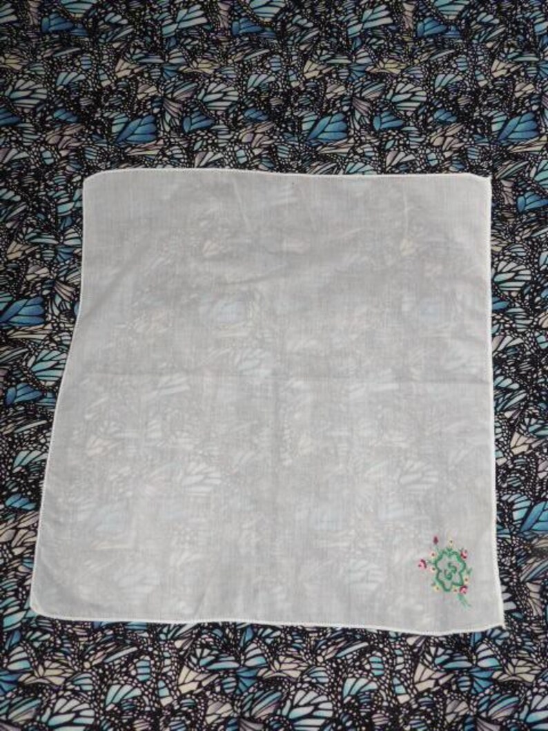 May include: A white cotton handkerchief with a small embroidered design of a green flower with red accents.