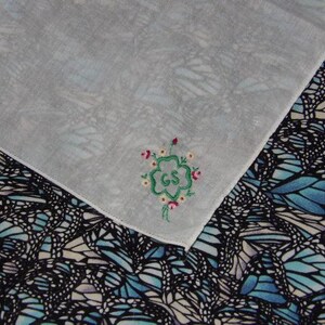 May include: White handkerchief with a green four-leaf clover embroidered with the letters "GS" in the center. The clover is surrounded by small pink flowers. The handkerchief is on top of a fabric with a blue and black butterfly pattern.