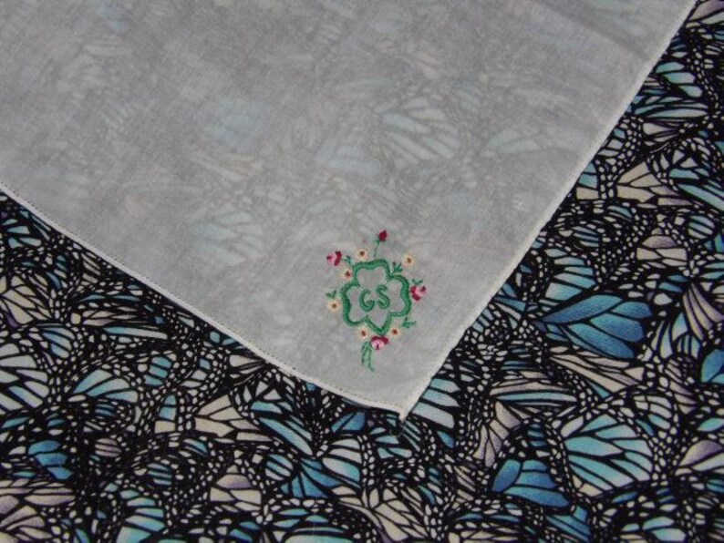 May include: White handkerchief with a green four-leaf clover embroidered with the letters "GS" in the center. The clover is surrounded by small pink flowers. The handkerchief is on top of a fabric with a blue and black butterfly pattern.
