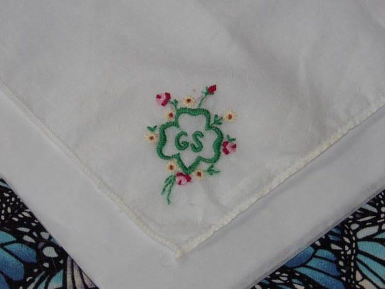 May include: White cotton handkerchief with green embroidered four-leaf clover with the letters "GS" in the center. The clover is surrounded by pink and white embroidered flowers.