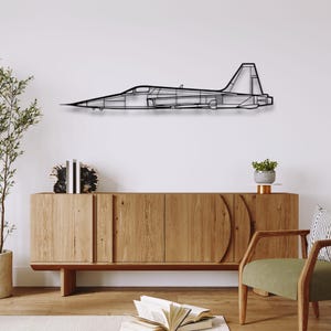 Puede incluir: Una pieza de arte mural de metal negro que representa un avión de combate de perfil. La obra de arte está montada en una pared blanca sobre un mueble de madera. El diseño del avión es minimalista, con contornos visibles del fuselaje, las alas y la cola.