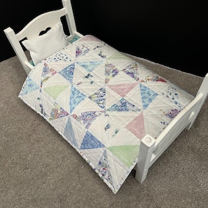 May include: A white wooden doll bed featuring a heart-shaped cutout on the headboard, a small white pillow, and a quilted blanket. The blanket is constructed from triangular fabric patches in shades of light blue, soft pink, green, and floral patterns.