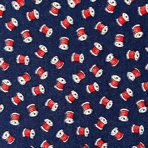 May include: A dark navy blue fabric featuring a repeating pattern of small, stylized sewing thread spools. Each spool is depicted with a vibrant red center representing the wound thread and white ends, arranged in a consistent, diagonal orientation.