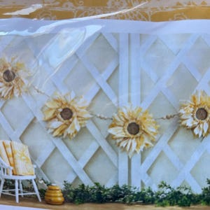 May include: A decorative sunflower garland with a string of paper flowers, each with a brown center and yellow petals. The garland hangs against a white lattice backdrop, with a small white chair and a decorative beehive.
