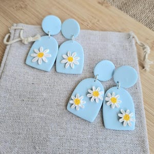 Könnte beinhalten: Hellblaue Gänseblümchen-Ohrringe. Jeder Ohrring hat eine hellblaue Bogenform mit einem weißen und gelben Gänseblümchen-Design. Die Ohrringe haben einen kleinen, runden hellblauen Stecker oben.