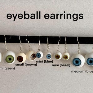 May include: A collection of eyeball earrings in various sizes and colors, including blue, green, brown, and hazel. The earrings are displayed on a black bar against a white background. The text "eyeball earrings" is at the top.