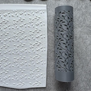 Puede incluir: Una hoja blanca rectangular texturizada con un patrón de hojas en relieve, junto a un objeto cilíndrico gris con un diseño de hojas a juego recortado. Los objetos están sobre una superficie gris.