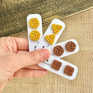 May include: Three sets of polymer clay earrings. The first set is yellow and features a swirl design. The second set is yellow and features a knotted triangle design. The third set is brown and features a swirl design and a knotted square design.