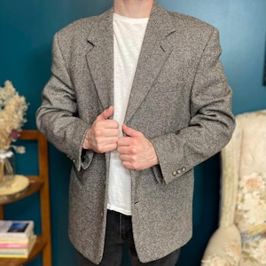 Op de afbeelding: Een grijs en bruin tweed blazer met een klassieke revers en een sluiting met drie knopen. De jas heeft een subtiel textuurpatroon en wordt gedragen over een wit t-shirt. De blazer is een vintage stijl.