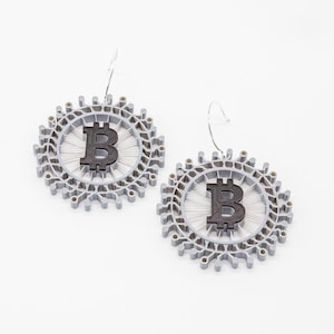 Könnte beinhalten: Ein Paar silberne Ohrringe mit einem schwarzen Bitcoin-Symbol in der Mitte jedes Ohrrings.