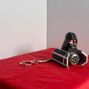 Puede incluir: Un llavero de Darth Vader negro y plateado, con una réplica detallada de su casco y caza TIE. El llavero tiene un cierre plateado y está sobre una superficie de terciopelo rojo.