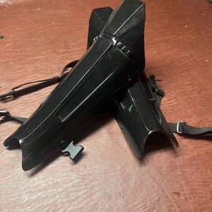 May include: Two black shin guards with adjustable straps and buckles. The guards have a glossy finish and are designed to protect the lower legs. They are made of a durable material and are likely for sports or cosplay.