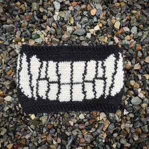 May include: A black and white crochet face covering with a toothy grin design. The handmade accessory features a black border and a white, open-mouth pattern, creating a unique and playful look. The item is likely a novelty item or costume piece.