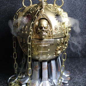 Puede incluir: Un quemador de incienso esférico dorado con detalles de calaveras y engranajes. El quemador tiene una cadena y se asienta sobre una base metálica. El humo emana de la parte superior y los lados, creando una atmósfera mística.