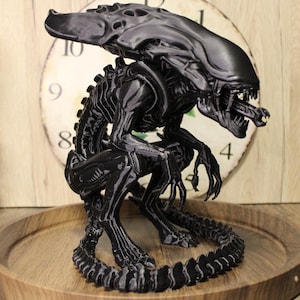 May include: A black Xenomorph figurine from the Alien franchise, featuring an elongated head, sharp teeth, a secondary inner jaw, and a long, segmented tail. The creature is depicted in a crouching, predatory stance with clawed hands and feet.