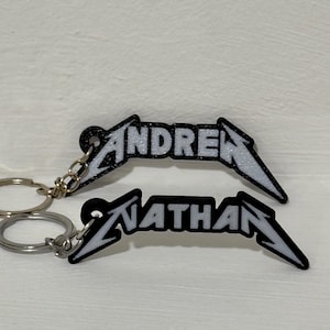Może przedstawiać: Dwa czarne metalowe breloki z imionami "ANDREW" i "NATHAN" w białych literach. Breloki są przymocowane do srebrnych kółek.