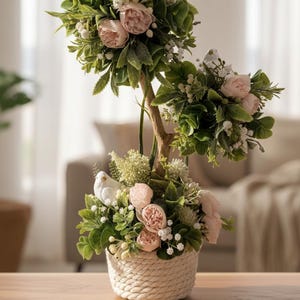 Puede incluir: Arreglo floral decorativo en una cesta tejida. El arreglo presenta rosas rosas, flores blancas y follaje verde, que se asemeja a un árbol en miniatura. La base es una cesta tejida, colocada sobre una superficie de madera.