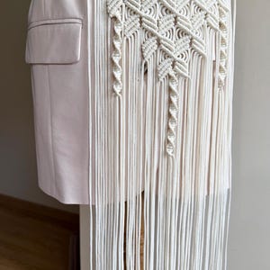 May include: Cream-colored macrame belt with a geometric design and long fringe. The belt is worn over a light pink blazer, adding a bohemian touch to the outfit. The intricate knotting creates a textured, eye-catching accessory.