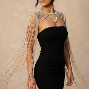 Puede incluir: Un vestido midi negro sin tirantes combinado con un accesorio decorativo dorado para los hombros. El accesorio presenta un diseño de cuello alto con largos flecos metálicos en cascada que caen por los brazos y un colgante central dorado.