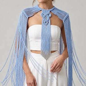 May include: A light blue macrame capelet featuring intricate knotted cord patterns, a high neckline, and long, flowing fringe that extends down the arms and chest. The design incorporates metallic silver cylindrical accents.