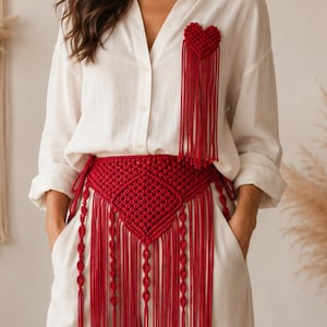 Op de afbeelding: Een wit overhemd met knopen en een bijpassende broek, gecombineerd met een rode macramé riem met een ruitpatroon en lange franjes, plus een bijpassende rode, hartvormige macramé broche op het borstgedeelte.