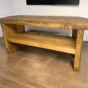 May include: A rustic wooden TV stand with a single shelf. The stand is made of natural wood with a warm brown finish. It has a simple, rectangular design with a slightly angled top.