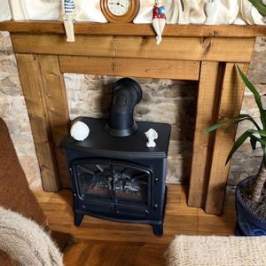 May include: A black electric fireplace with a curved flue is set within a wooden mantel. The fireplace has a glass door and a decorative interior. A small white sculpture sits on top of the fireplace.