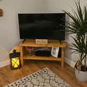 May include: A corner TV stand made of reclaimed wood with a dark wood finish. The stand has two shelves and is holding a flat screen TV, a decorative box with the text "Live Happy", a small plant, and a framed photo. The stand is in a living room with a rug on the floor and a potted plant in the corner.