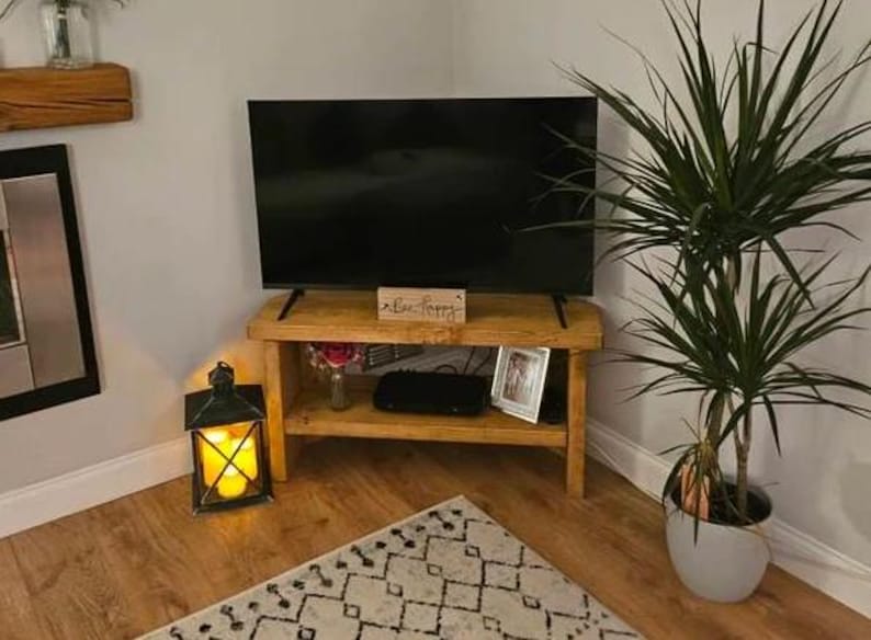 May include: A corner TV stand made of reclaimed wood with a dark wood finish. The stand has two shelves and is holding a flat screen TV, a decorative box with the text "Live Happy", a small plant, and a framed photo. The stand is in a living room with a rug on the floor and a potted plant in the corner.