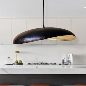 Könnte beinhalten: Eine moderne Pendelleuchte in Schwarz und Gold. Die Leuchte hat eine strukturierte, gehämmert Metallaußenseite und eine goldene Innenseite. Sie hängt an einem schwarzen Kabel in einer Küche. Eine stilvolle Ergänzung für jedes Zuhause.